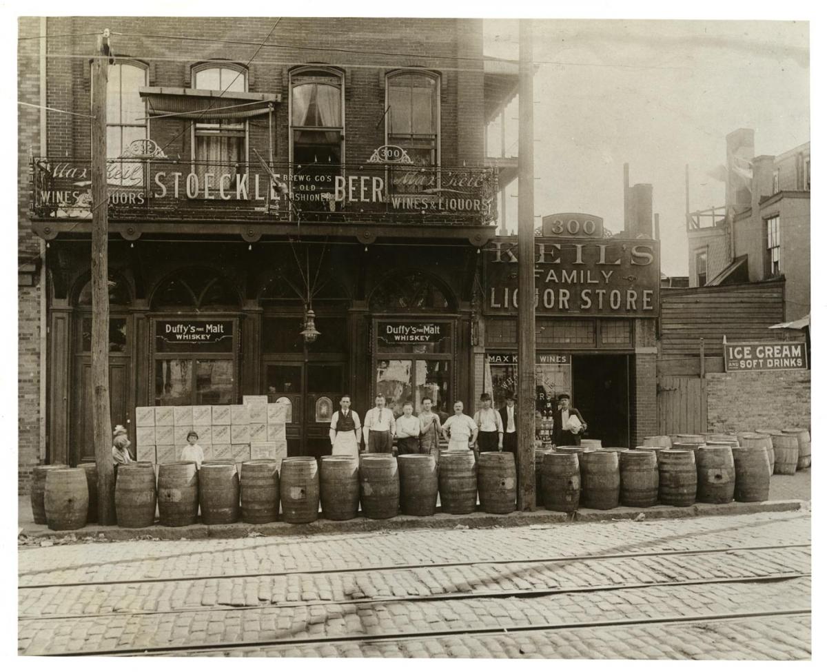 Men stand outside a tavern and liquor store with barrels lining the sidewalk