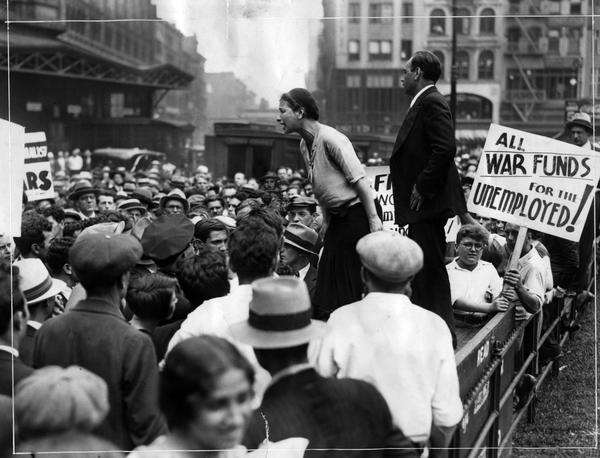 Photograph of a protest. June Croll is speaking to the crowd of people. There is a sign in front of her that reads, "All war funds for the unemployed."