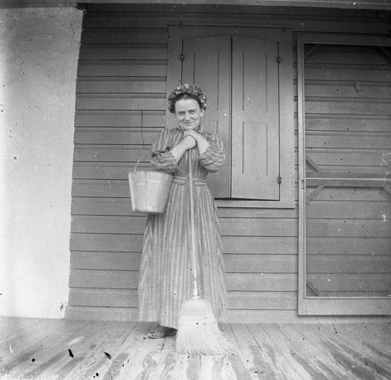 Unidentified woman with broom and bucket on porch