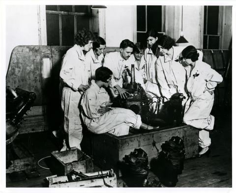 Group of women in flights suits gathered around two men demonstrating the parts of an aircraft motor.