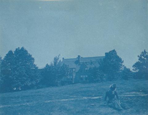 Cyanotype photo of a person sitting in a grassy field with trees and a large house in the background.