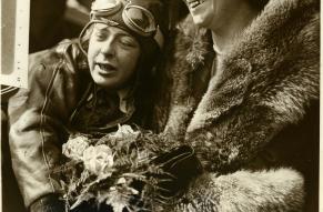 Black and white photograph of a woman in a pilot's gear hugging a woman holding flowers.