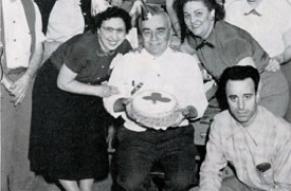 A black and white photograph depicts a group of people celebrating St. Patrick's Day, with a man holding a cake.