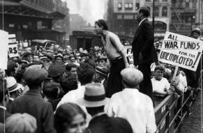 Photograph of a protest. June Croll is speaking to the crowd of people. There is a sign in front of her that reads, "All war funds for the unemployed."